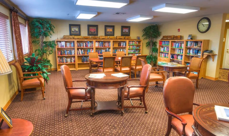 A cozy senior living facility library room with wooden bookshelves filled with books along the back wall, several round wooden tables surrounded by cushioned chairs, potted plants in the corners, framed pictures on the wall, and a clock above a doorway.