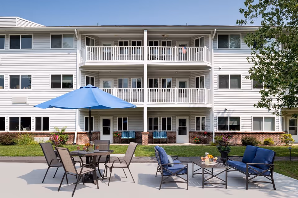 Outdoor patio area in front of a three-story senior living facility building with white siding and brick accents. The patio features a round table with four chairs and a large blue umbrella, as well as a seating area with blue cushioned chairs and a small table with drinks and flowers. Green grass and some plants surround the patio.
