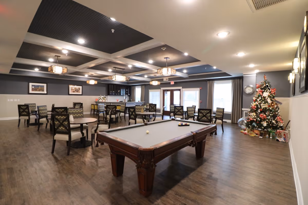 A spacious common room with a pool table in the foreground, several round tables with chairs arranged around the room, and a decorated Christmas tree in the corner. The room features a coffered ceiling with hanging light fixtures, framed pictures on the walls, and large windows with curtains allowing natural light to enter.