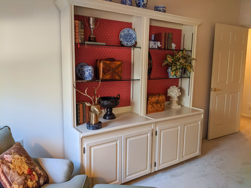 A cozy living room corner featuring a built-in white shelving unit with glass shelves displaying decorative items such as blue and white ceramics, books, a brass deer head sculpture, a plant, and a small chest. To the left is a cushioned armchair with a floral pillow, and to the right is an open door leading to another room.