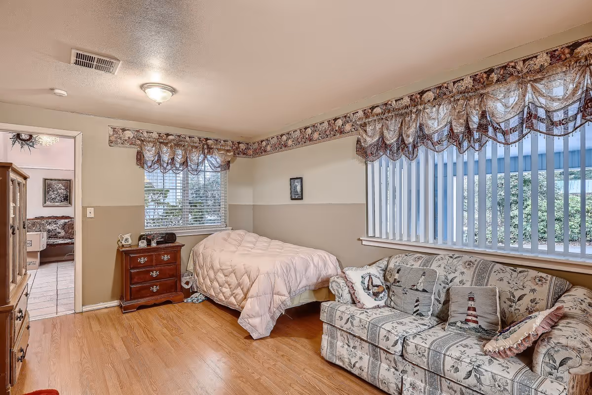A cozy bedroom with a single bed covered in a light pink comforter, a wooden nightstand with a clock and decorative items, a floral-patterned couch with lighthouse-themed pillows, and large windows with vertical blinds and floral valances. The room has wood flooring and beige walls with a floral wallpaper border near the ceiling. An open doorway leads to another room with a floral sofa and tiled floor.