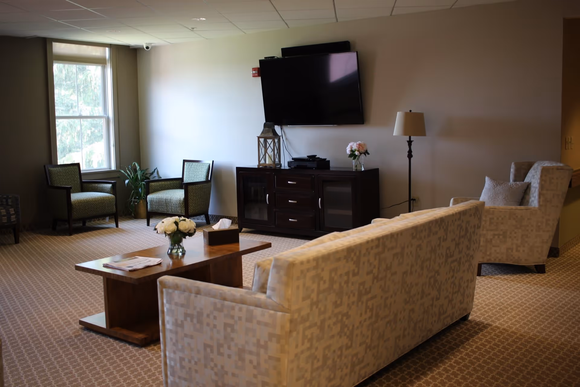 A furnished senior living common room with sofas and armchairs arranged around a wooden coffee table, a wall-mounted TV, and a floor lamp.