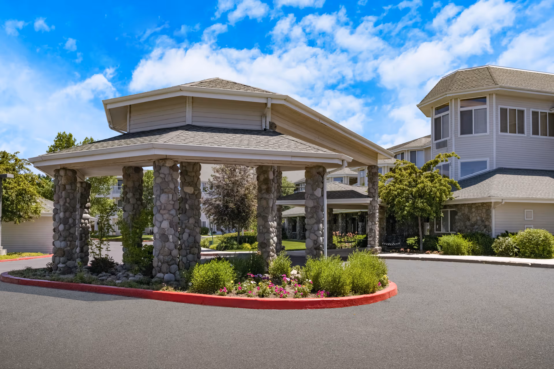 Entrance area of Stone Lodge Gracious Retirement Living featuring a covered driveway supported by stone pillars, surrounded by landscaped greenery and flowers, with the building visible in the background under a partly cloudy blue sky.