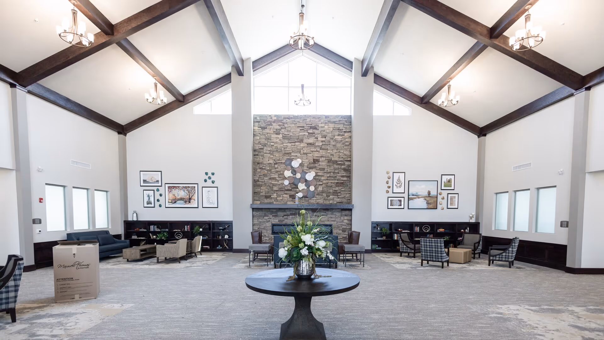 A spacious senior living facility common area with high vaulted ceilings and exposed wooden beams. The room features a large stone fireplace centered on the far wall, flanked by framed artwork and decorative items. Multiple seating areas with chairs and sofas are arranged around the room, and a round table with a floral arrangement is positioned in the foreground. The space is well-lit with natural light from windows and hanging chandeliers.