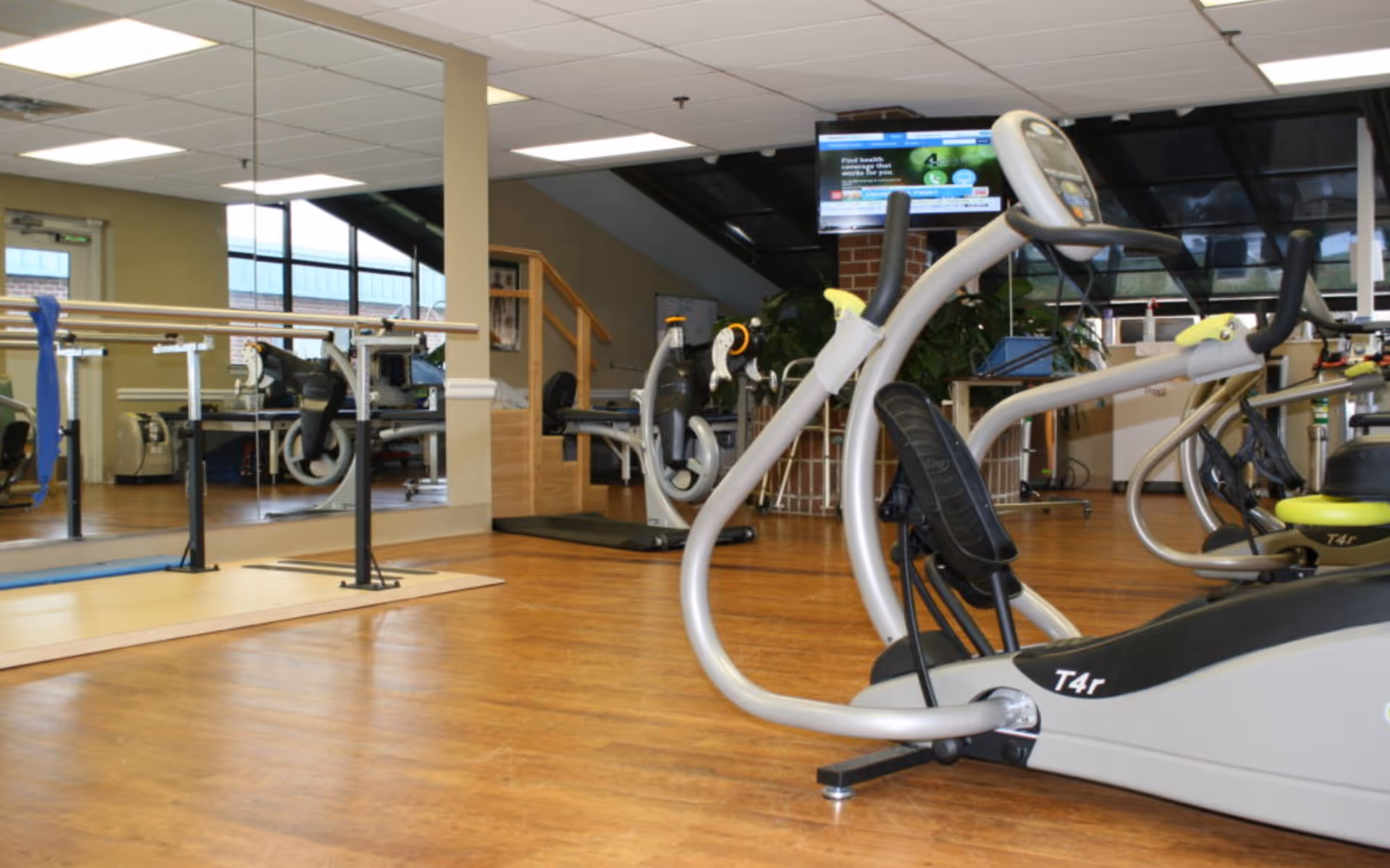 Interior view of a fitness room in a senior living facility featuring exercise equipment including stationary bikes and elliptical machines, wooden flooring, large mirrors on the wall, and a television mounted in the background.