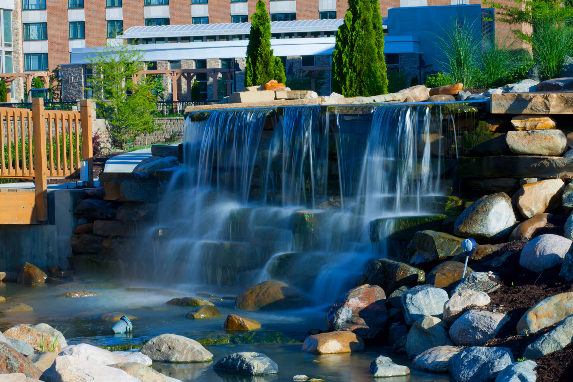 A serene outdoor waterfall feature with water cascading over rocks into a pond, surrounded by landscaping with trees and shrubs. In the background, there is a multi-story building with large windows and a wooden fence on the left side.