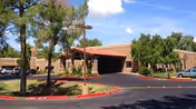Exterior view of a single-story assisted living facility building with a covered entrance, surrounded by trees and parked cars under a blue sky with some clouds.