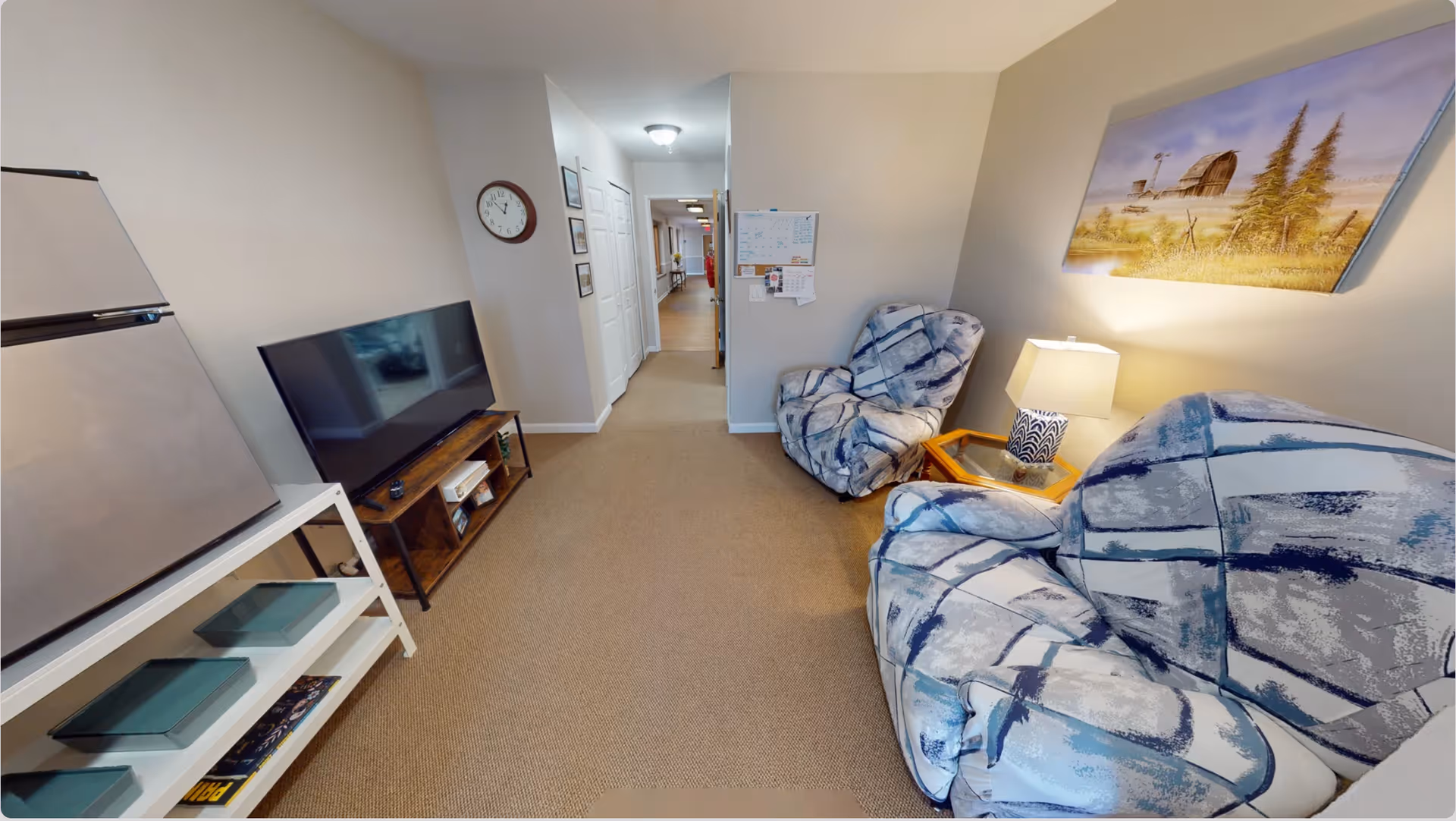 A cozy living room area in an assisted living facility featuring two patterned armchairs, a small side table with a lamp, a flat-screen TV on a wooden stand, a mini refrigerator, and a hallway leading to other rooms. A landscape painting hangs on the wall above one of the chairs, and a clock is mounted on the wall near the hallway.
