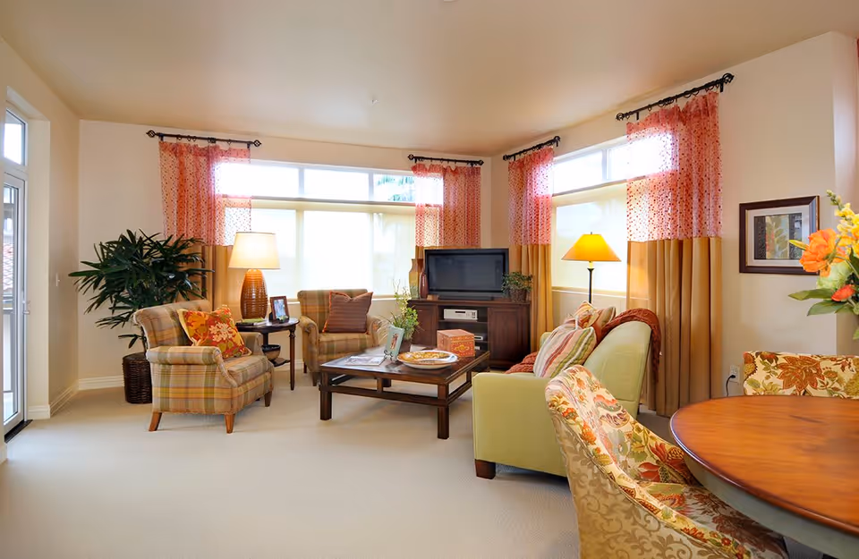 A bright and cozy living room with large windows covered by red and beige curtains. The room features a green sofa with patterned cushions, two plaid armchairs, a wooden coffee table with magazines and decorative items, a TV on a wooden stand, two table lamps, a potted plant, and a round wooden dining table with floral upholstered chairs.