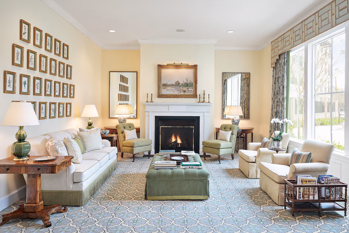 A bright and elegant living room with a lit fireplace centered on the far wall. The room features a white sofa with multiple cushions on the left, two green armchairs flanking the fireplace, and two beige armchairs on the right near a large window with patterned curtains. There are two side tables with lamps, a large green ottoman in the center with books on top, and framed botanical prints on the left wall. The floor is covered with a patterned carpet.