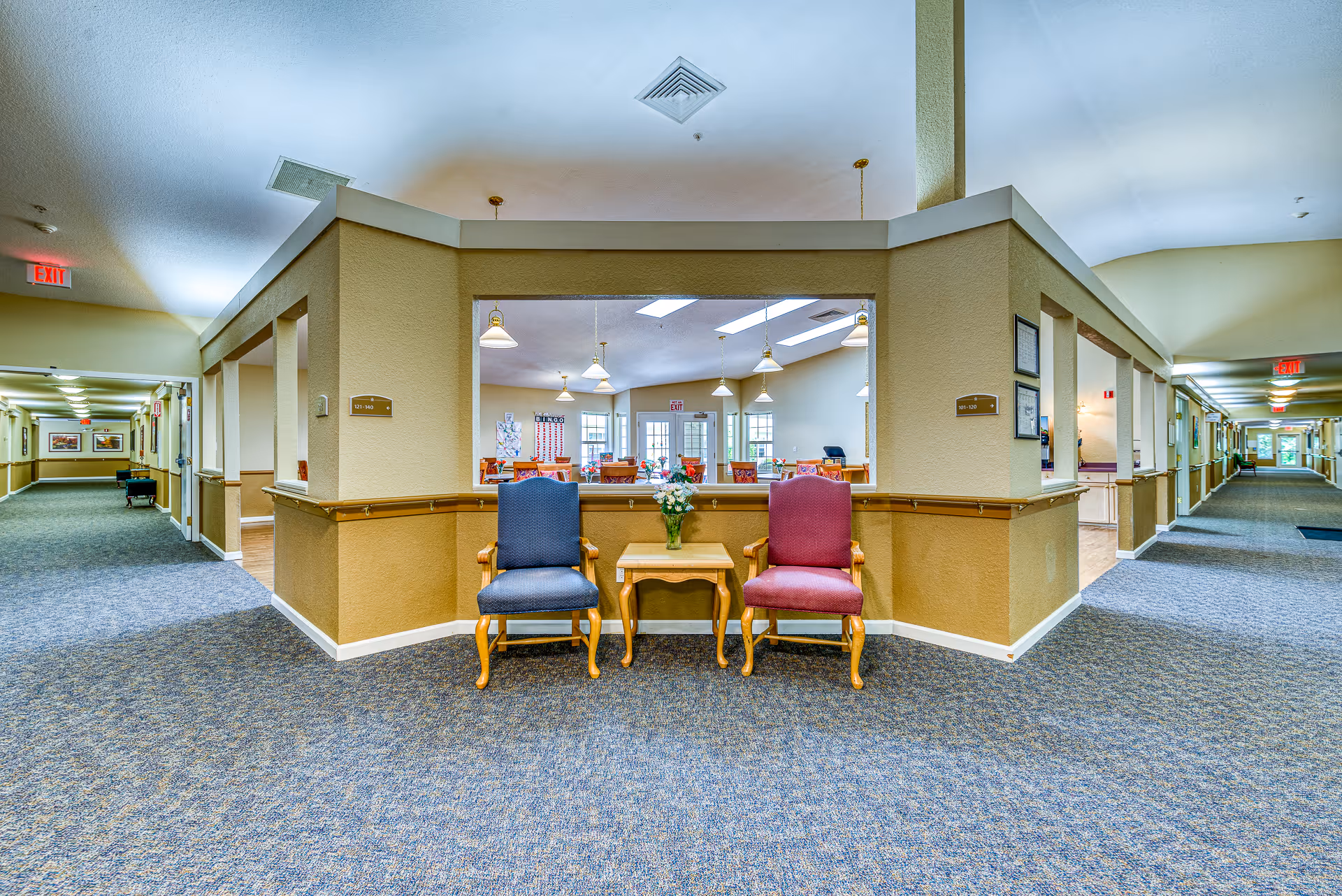 Interior view of a senior living facility hallway with two upholstered chairs, one blue and one red, placed on either side of a small wooden table with a vase of flowers. The hallway has beige walls with handrails and carpeted floors. Through a large window opening, a dining area with tables, chairs, hanging lights, and windows is visible.