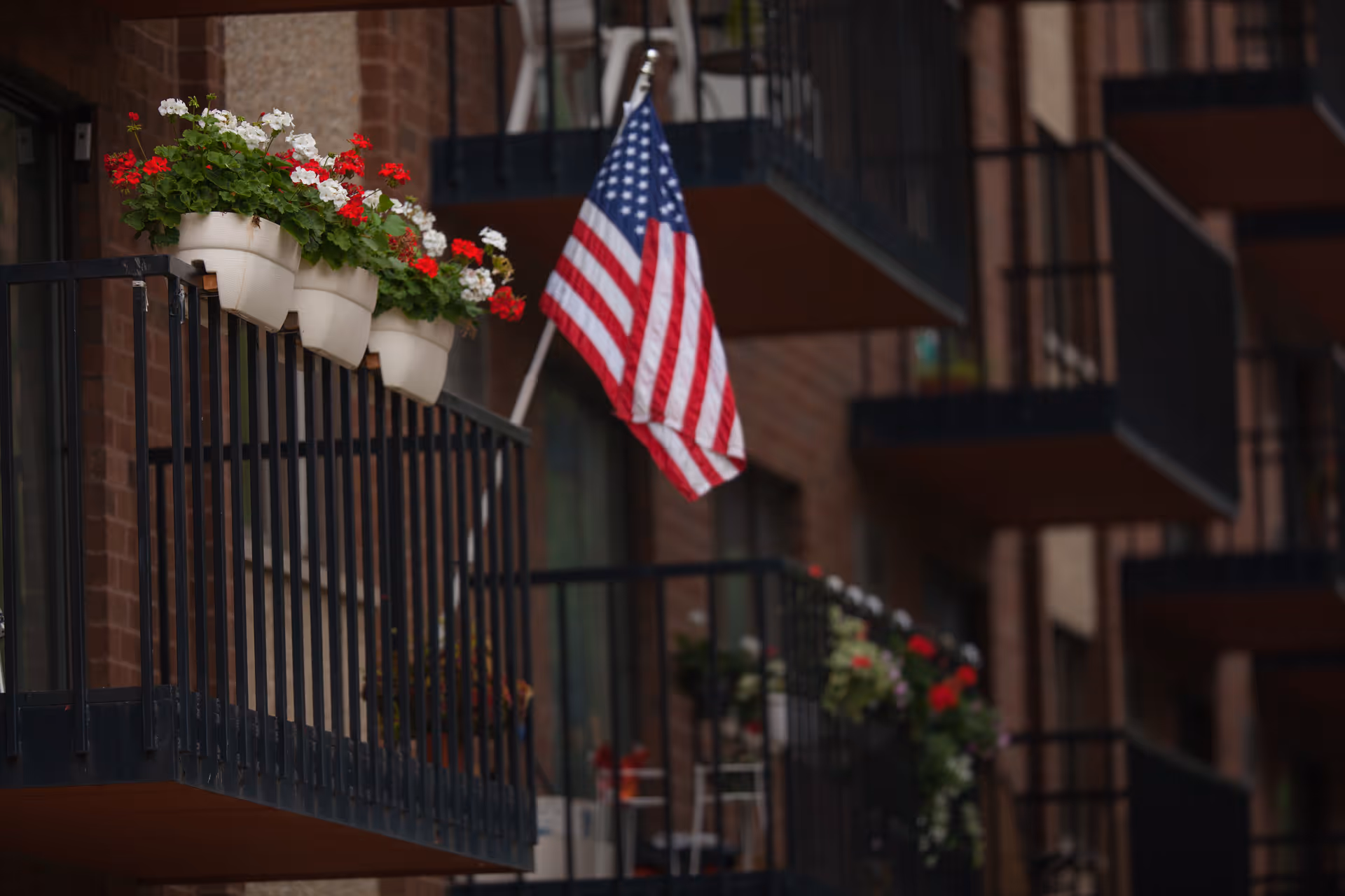 Close-up view of a balcony with flower pots containing red and white flowers and an American flag attached to the railing. Other balconies with similar railings and flower pots are visible in the background.