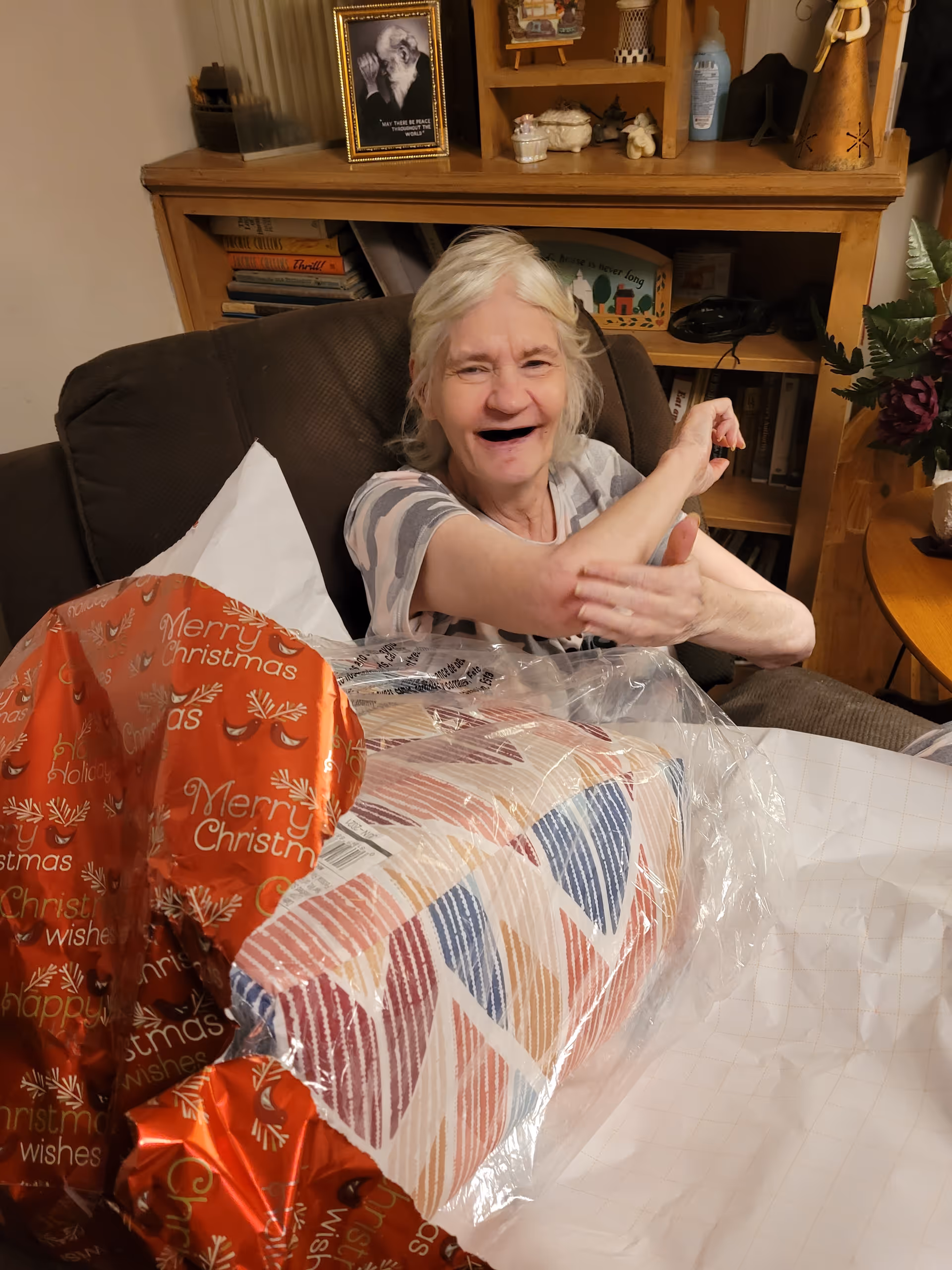 An elderly woman sitting on a brown armchair, smiling and gesturing with her arms. In front of her is a partially unwrapped gift with colorful patterned wrapping paper and red Christmas-themed wrapping paper. Behind her is a wooden bookshelf with various items including books, a framed photo, and decorative objects.