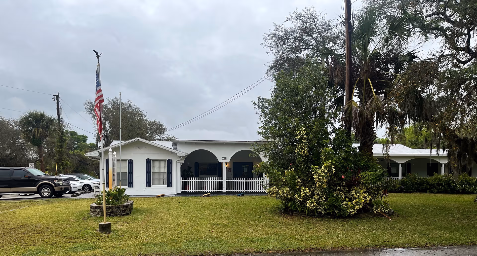 Front exterior view of a single-story white building with a porch and arched openings, surrounded by green grass and trees. An American flag is flying on a flagpole in the front yard, and several parked vehicles are visible on the left side.