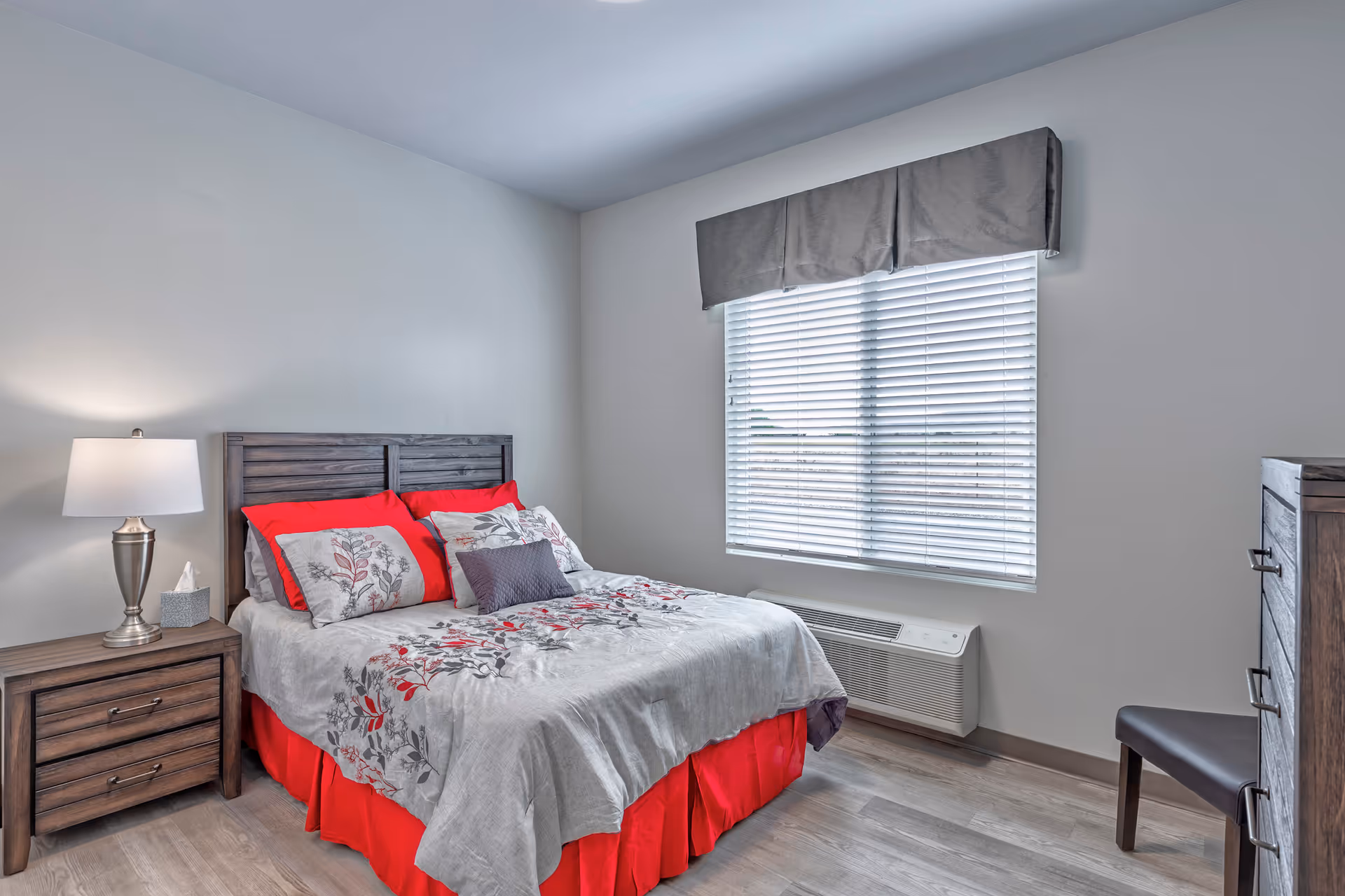 Bright bedroom with a made bed featuring red and gray bedding, a wooden nightstand with lamp, a window with blinds, dresser and chair.