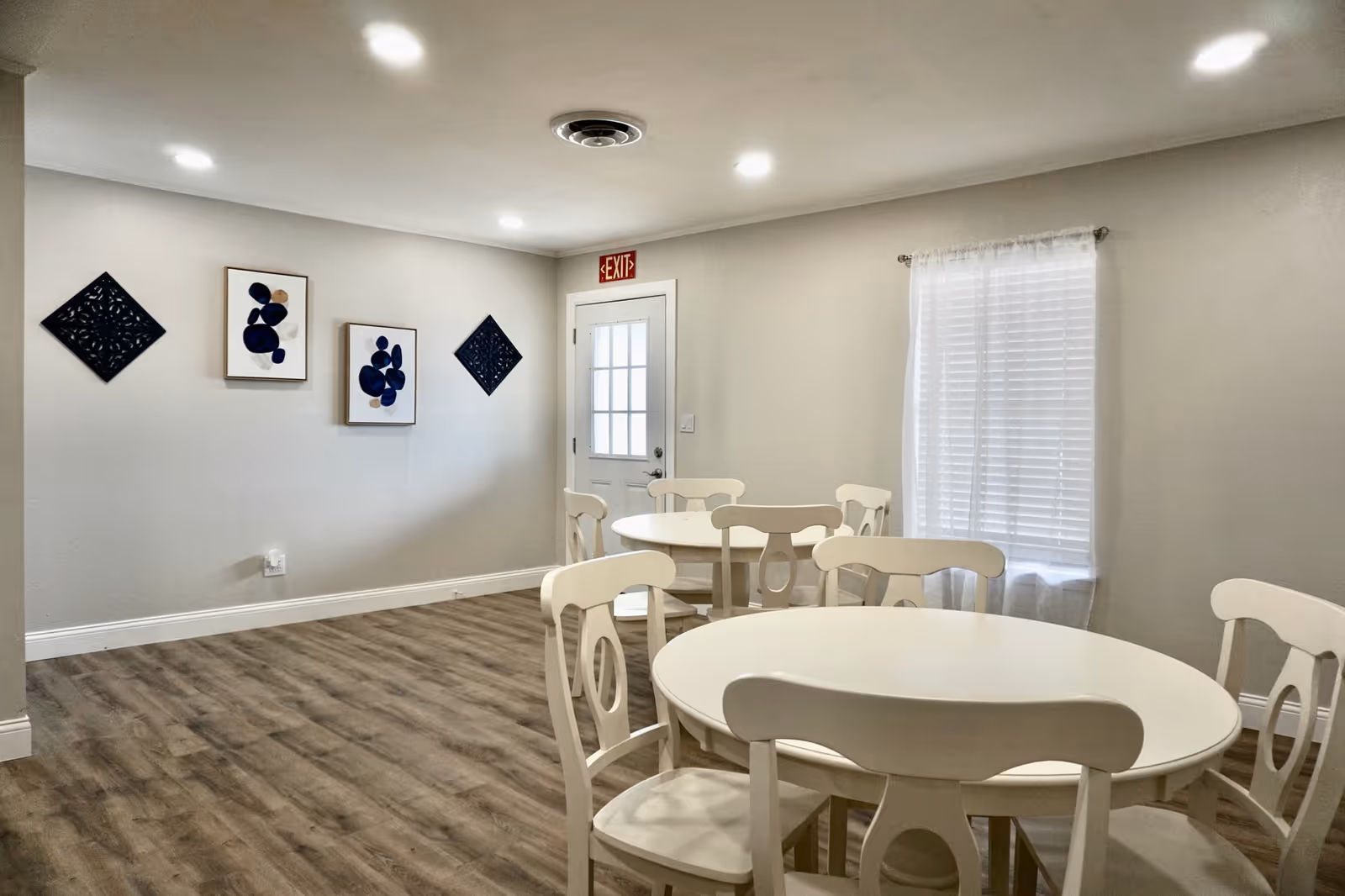 Communal dining area with round white tables and chairs, wood-look flooring, wall art, a window, and an exit door.