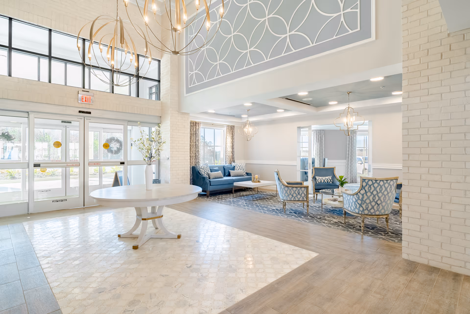 Bright and spacious senior living facility lobby with large windows, a round white table with a vase of flowers in the center, elegant chandeliers, and a seating area with blue upholstered chairs and a sofa on a patterned rug.