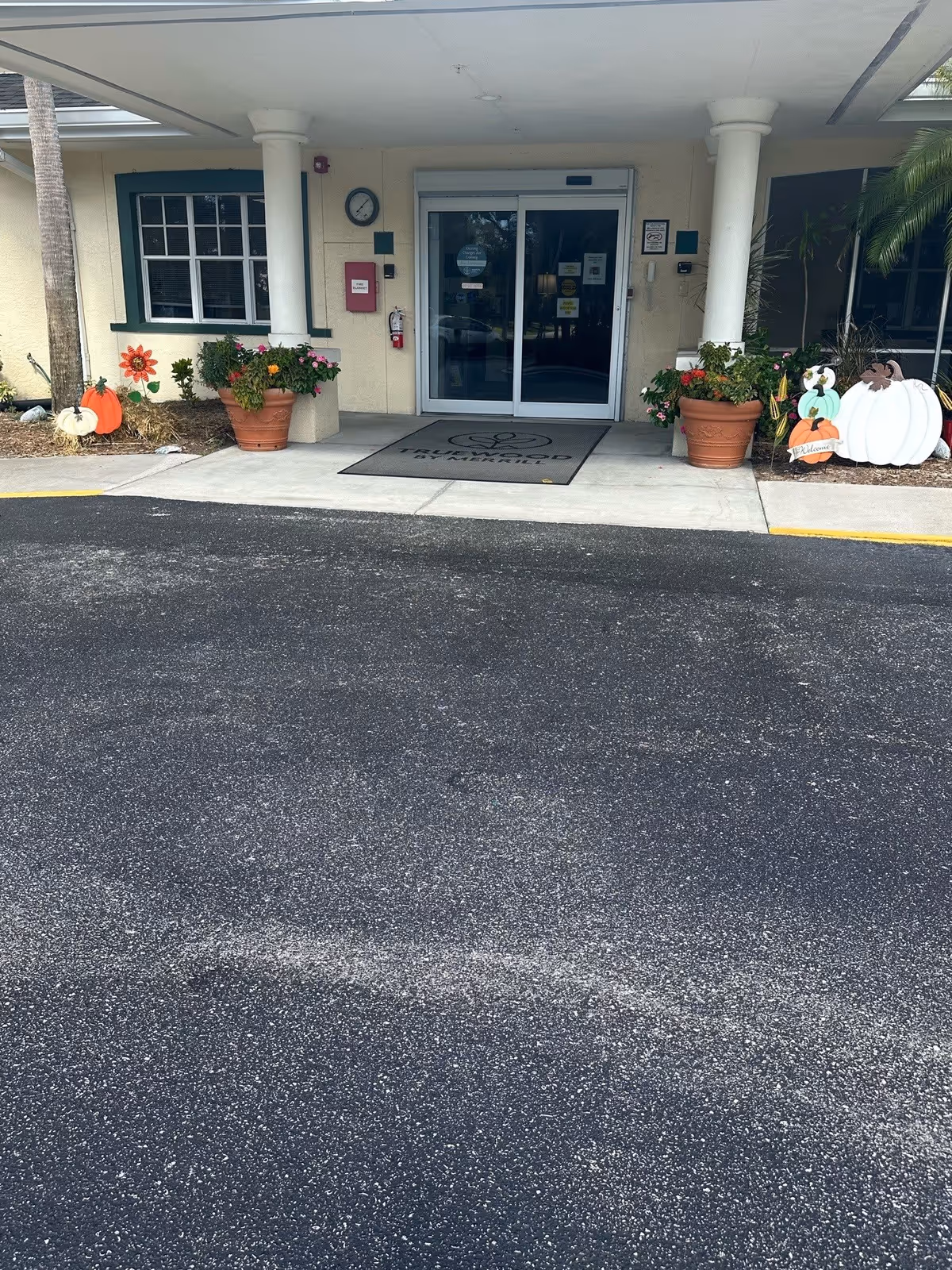 Entrance to Truewood by Merrill, Charlotte Center with automatic sliding glass doors, two white columns, potted plants on either side, and seasonal pumpkin decorations on the ground. There is a welcome mat with the facility's logo in front of the door.