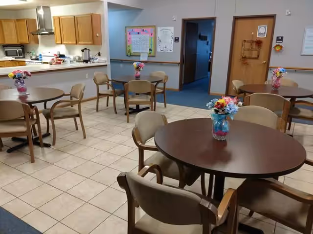 A dining area in a senior living facility with round wooden tables and beige cushioned chairs. Each table has a small vase with colorful artificial flowers. The room has tiled flooring and a kitchen area with wooden cabinets and appliances in the background. There are two doors and some wall decorations visible on the far wall.