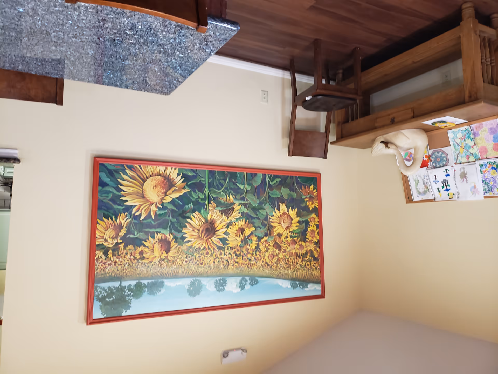 Interior room with a large framed painting of a sunflower field on a beige wall. Below the painting is a wooden chair and a wooden table with a swan sculpture and colorful drawings pinned on a corkboard. In the foreground, there is a granite countertop with wooden chairs around it.