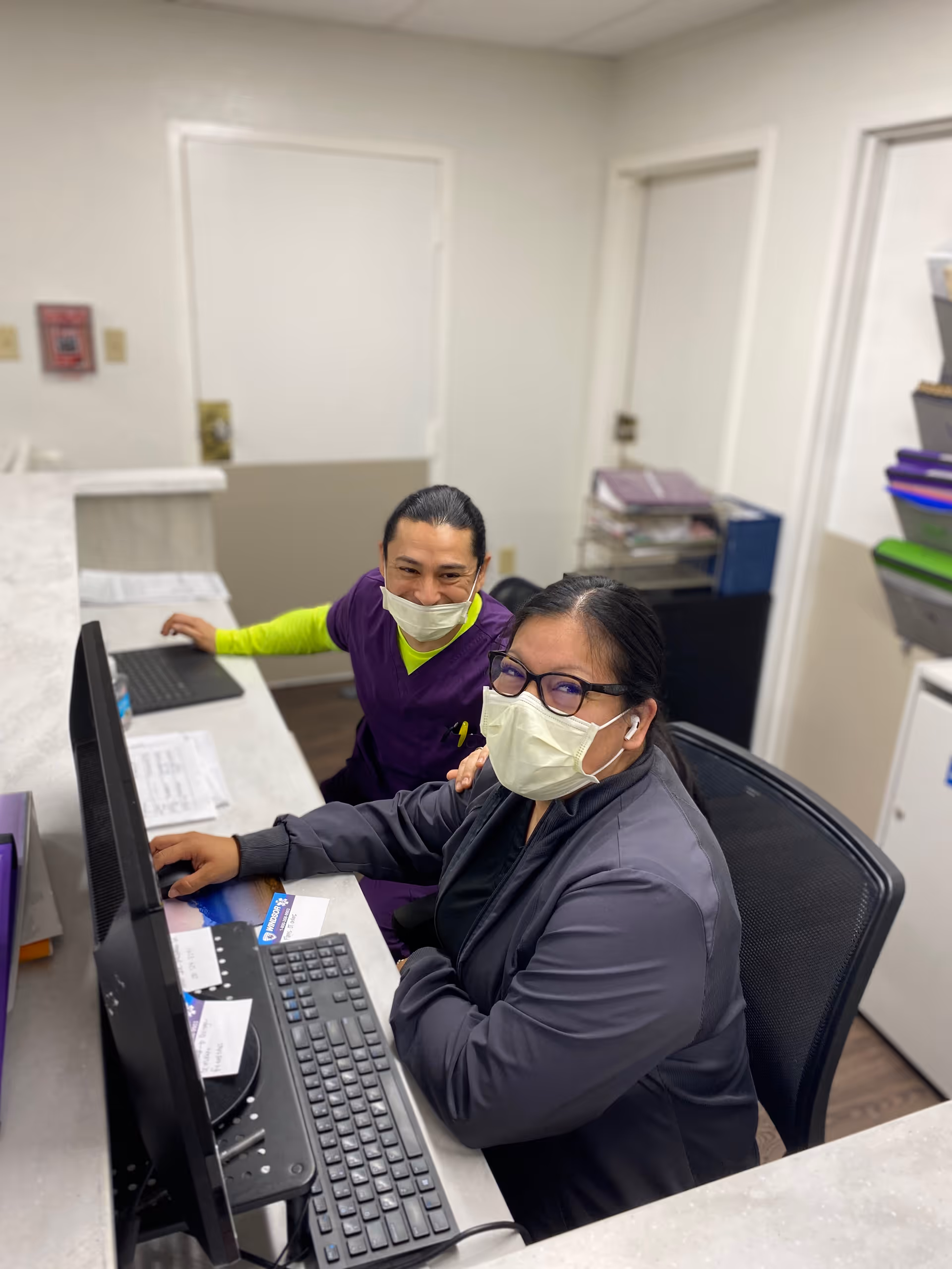 Two healthcare workers wearing masks are seated at a reception desk with a computer and keyboard. One is wearing a purple uniform with a neon green undershirt, and the other is wearing glasses and a dark jacket. They are smiling and looking at the camera in a well-lit office area with white walls and doors in the background.