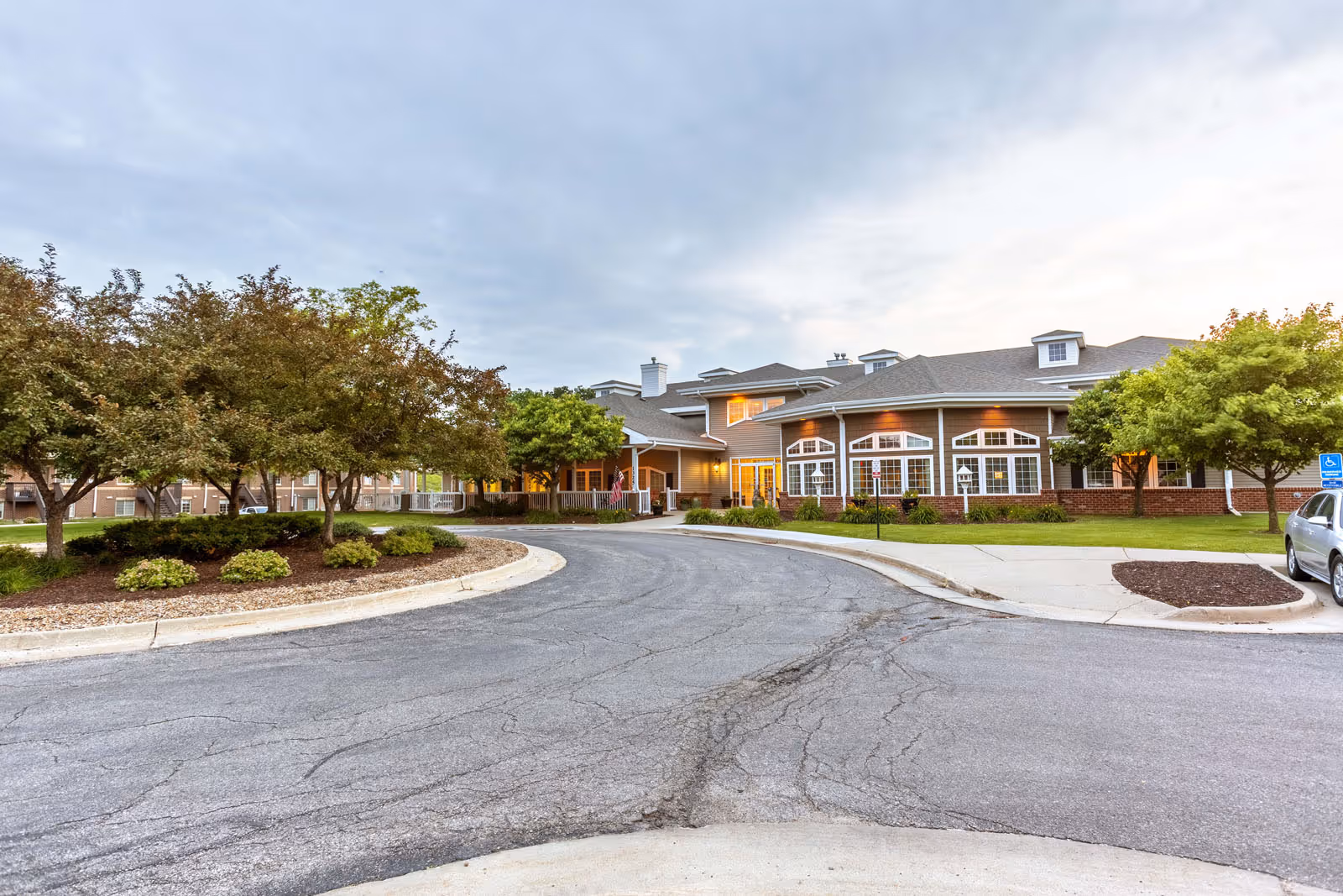 Front exterior of a senior living facility with a circular driveway, landscaped trees, and an illuminated entrance.
