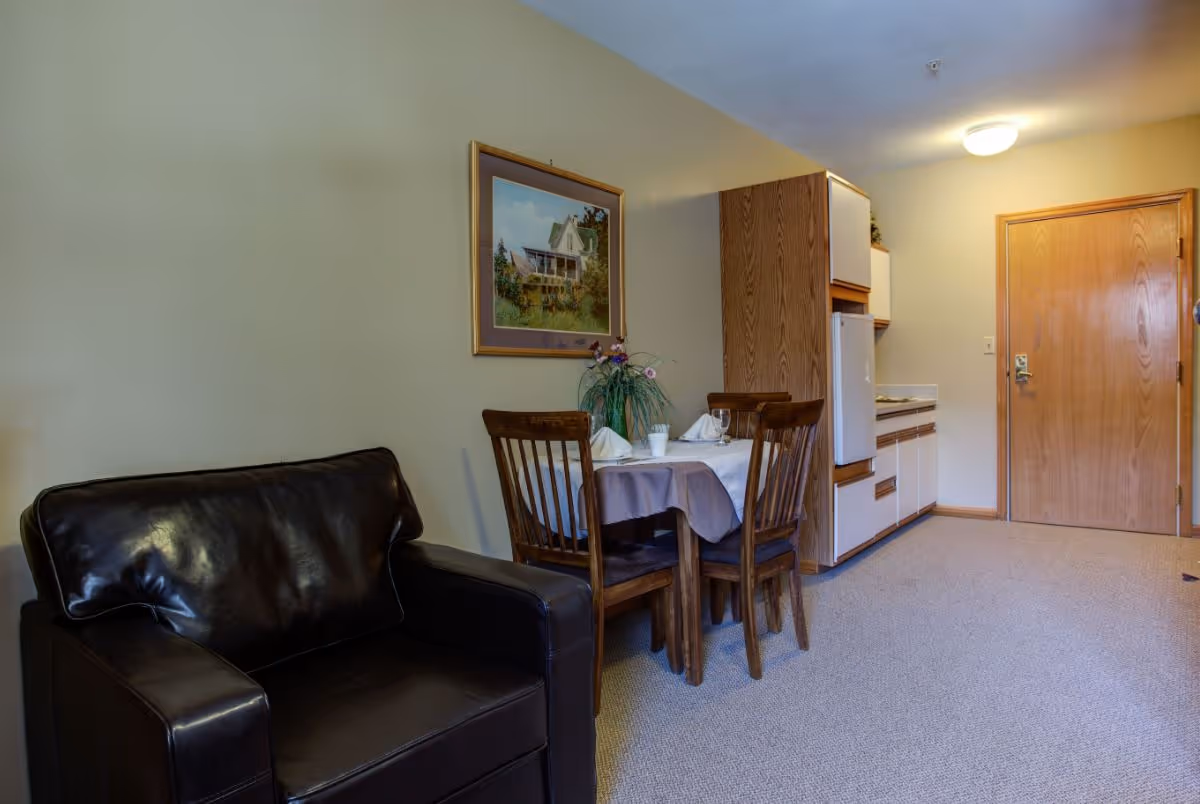 Small furnished interior with a leather armchair, a dining table set for two, a kitchenette, and an entry door.