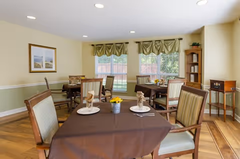 Small dining room with several tables set for meals, upholstered chairs, windows with green valances, and light wood floors.
