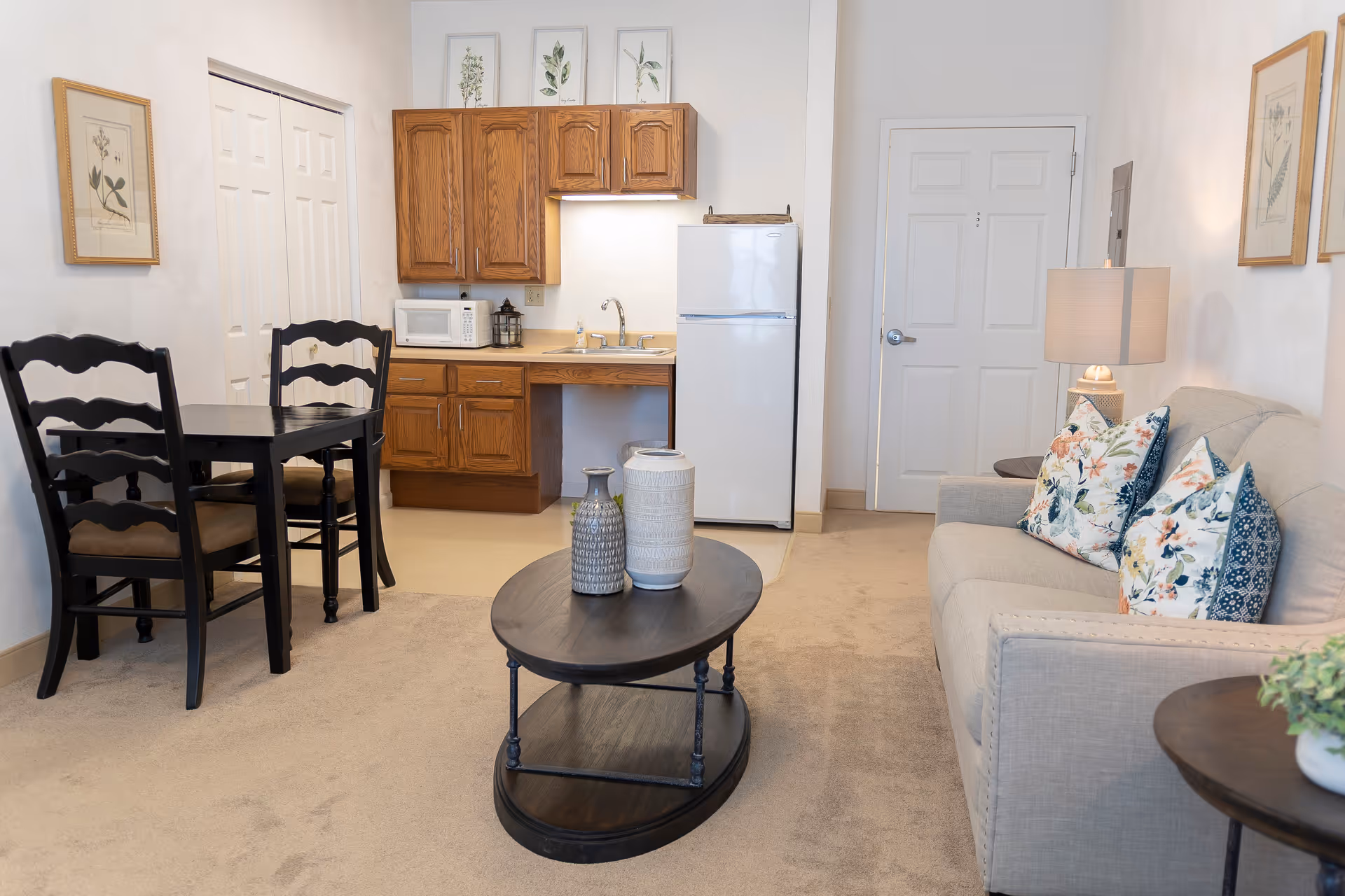 A cozy senior living space featuring a small kitchen area with wooden cabinets, a white refrigerator, a microwave, and a sink. Adjacent to the kitchen is a black dining table with two chairs. The living area includes a beige sofa adorned with floral and patterned pillows, a dark wooden coffee table with decorative vases, a side table with a lamp, and framed botanical artwork on the walls.