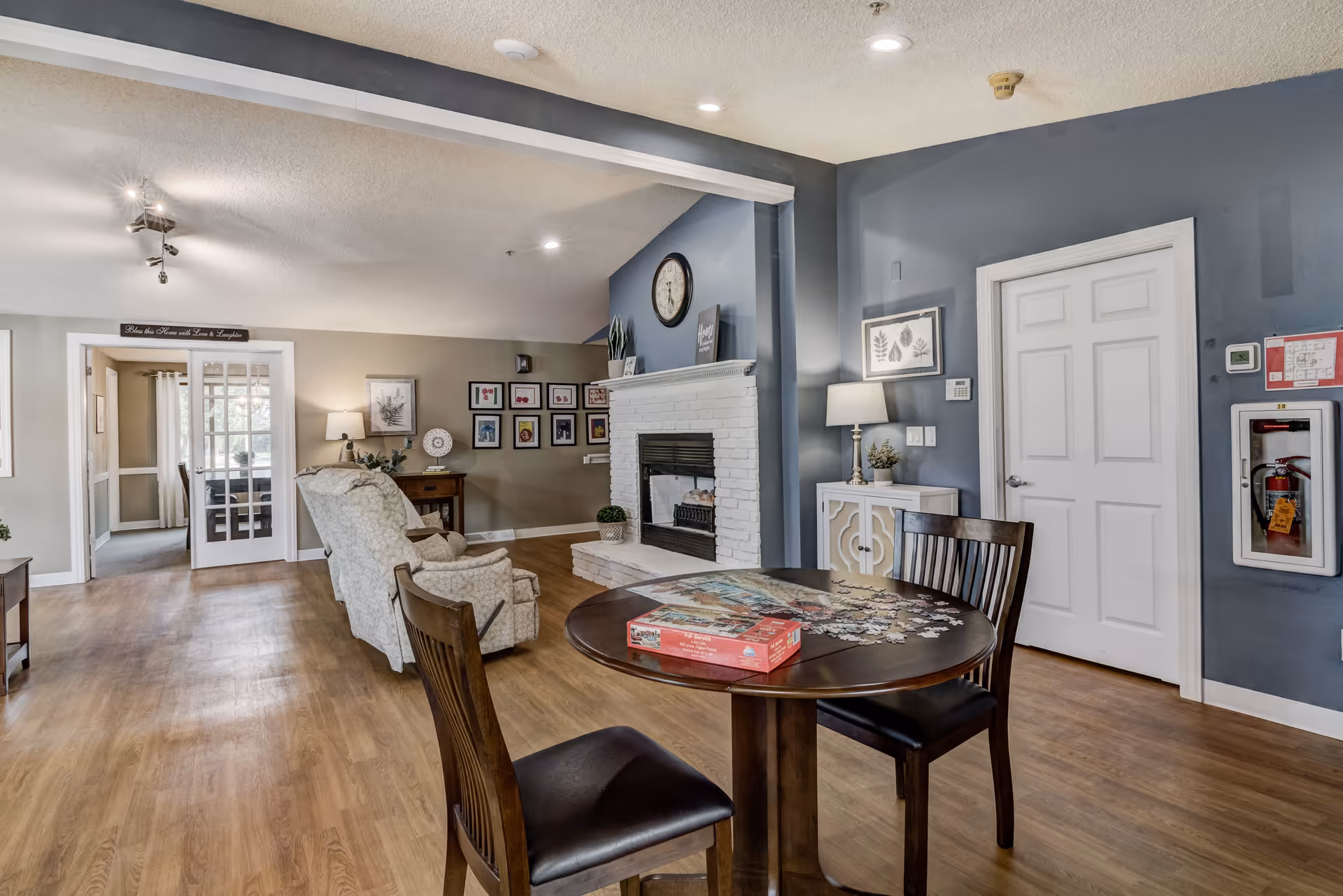 A cozy living room area with a round wooden table and two chairs in the foreground, a partially completed puzzle on the table, and a box for the puzzle. In the background, there are two upholstered armchairs facing a white brick fireplace with a clock above it. The walls are painted in shades of blue and beige, decorated with framed pictures and a lamp on a small cabinet. A door and a fire extinguisher are visible on the right side, and a hallway with glass double doors is seen on the left.