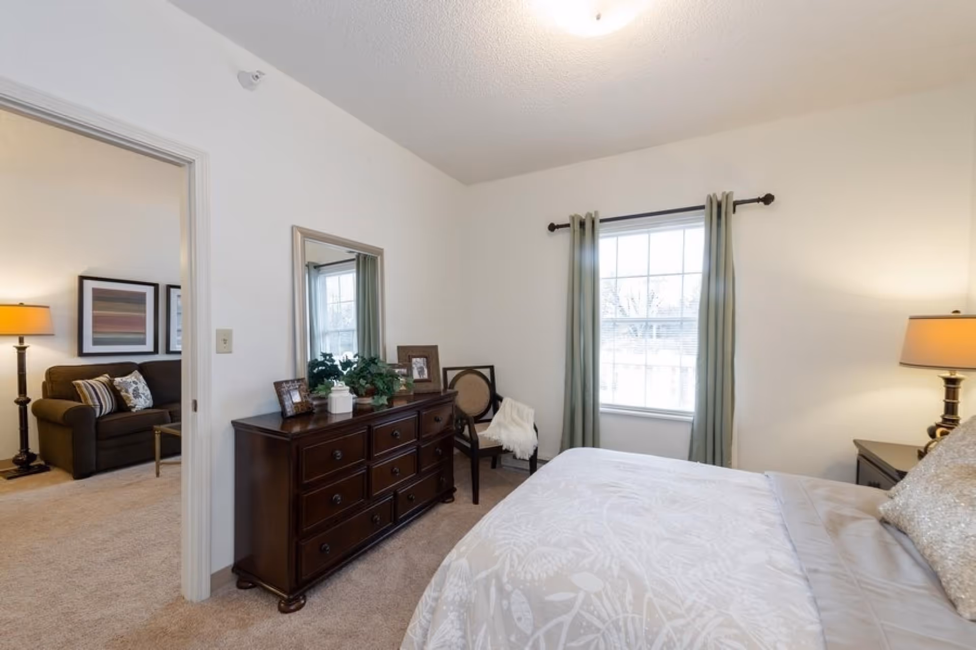 Bright furnished bedroom with a bed, dark wood dresser topped with decor and a mirror, a window with green curtains, and a glimpse into a connected sitting area.
