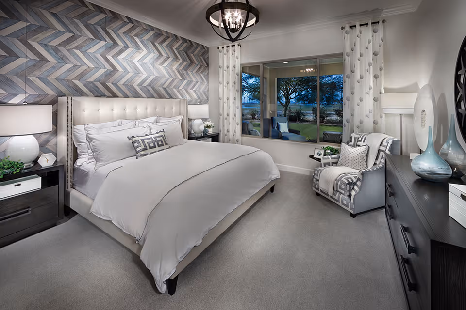 A modern bedroom with a large upholstered bed featuring white bedding and decorative pillows. The wall behind the bed has a geometric patterned accent wall in shades of gray and blue. There are two bedside tables with lamps, a comfortable armchair with a throw blanket and pillow, a dark dresser with decorative vases, and a large window showing an outdoor view with trees and a patio area. The room is carpeted and has a contemporary chandelier hanging from the ceiling.