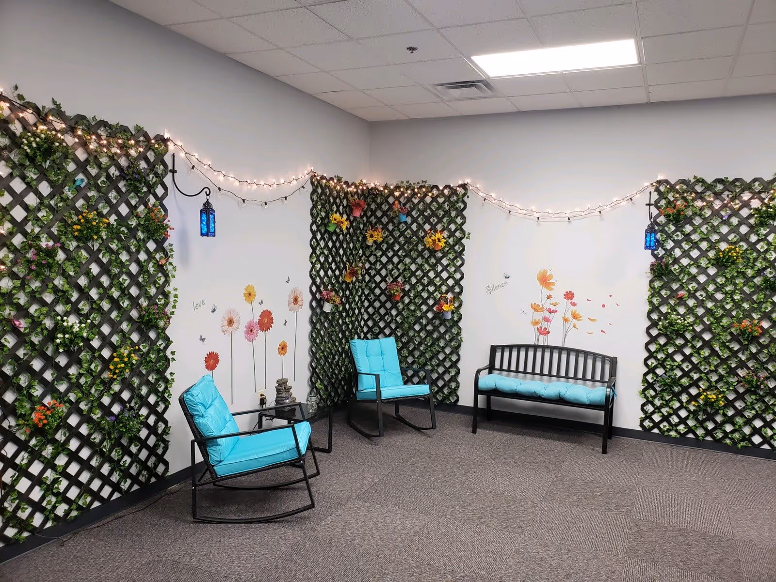 Indoor lounge area with turquoise-cushioned chairs and a bench against walls decorated with lattice panels, string lights, and flower decals.