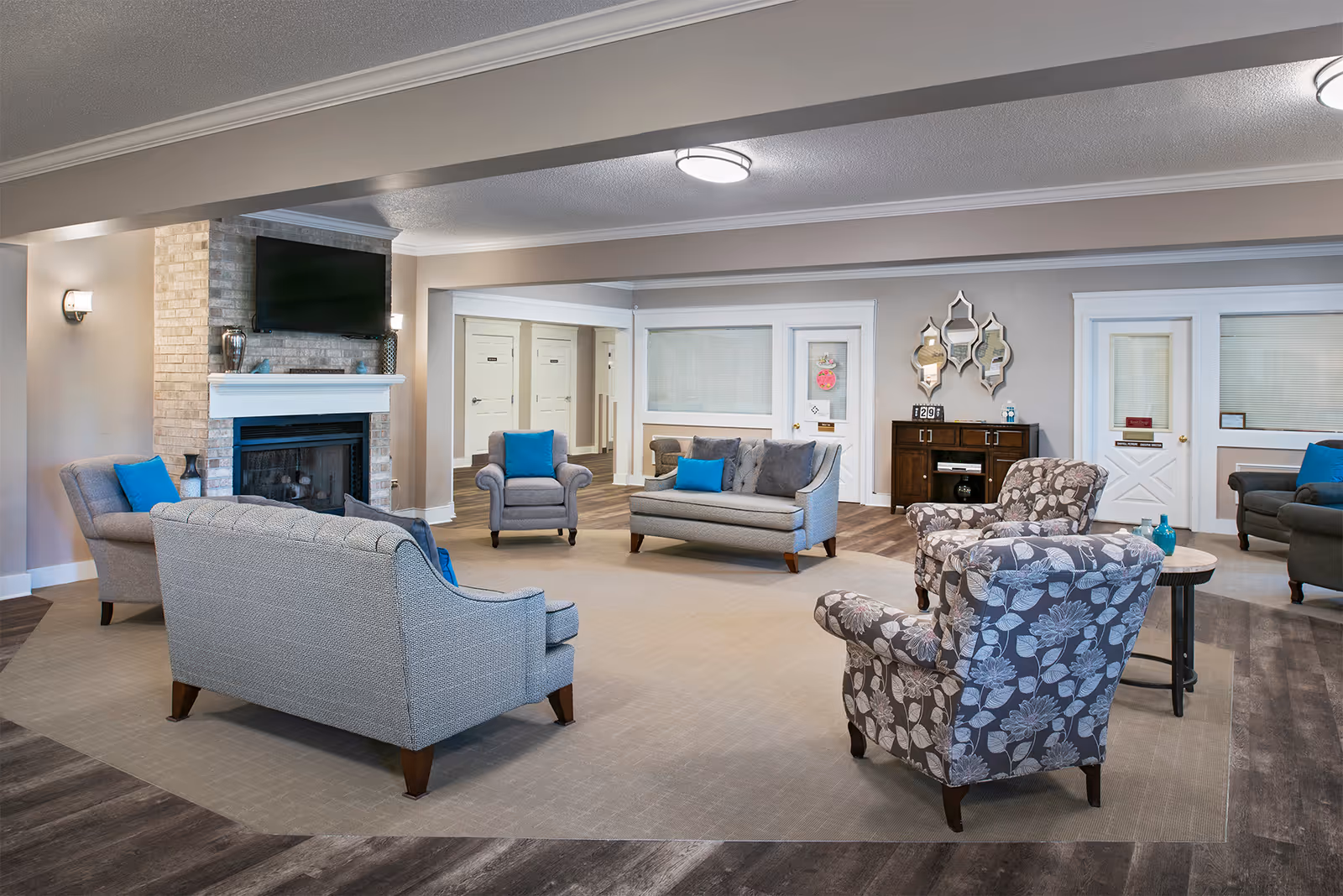 A spacious and well-lit living room area in a senior living facility featuring multiple upholstered chairs and sofas arranged around a central carpeted area. There is a brick fireplace with a mounted flat-screen TV above it, and a wooden cabinet with decorative mirrors on the wall. The room has neutral-colored walls and wood flooring, with blue accent pillows on the furniture.