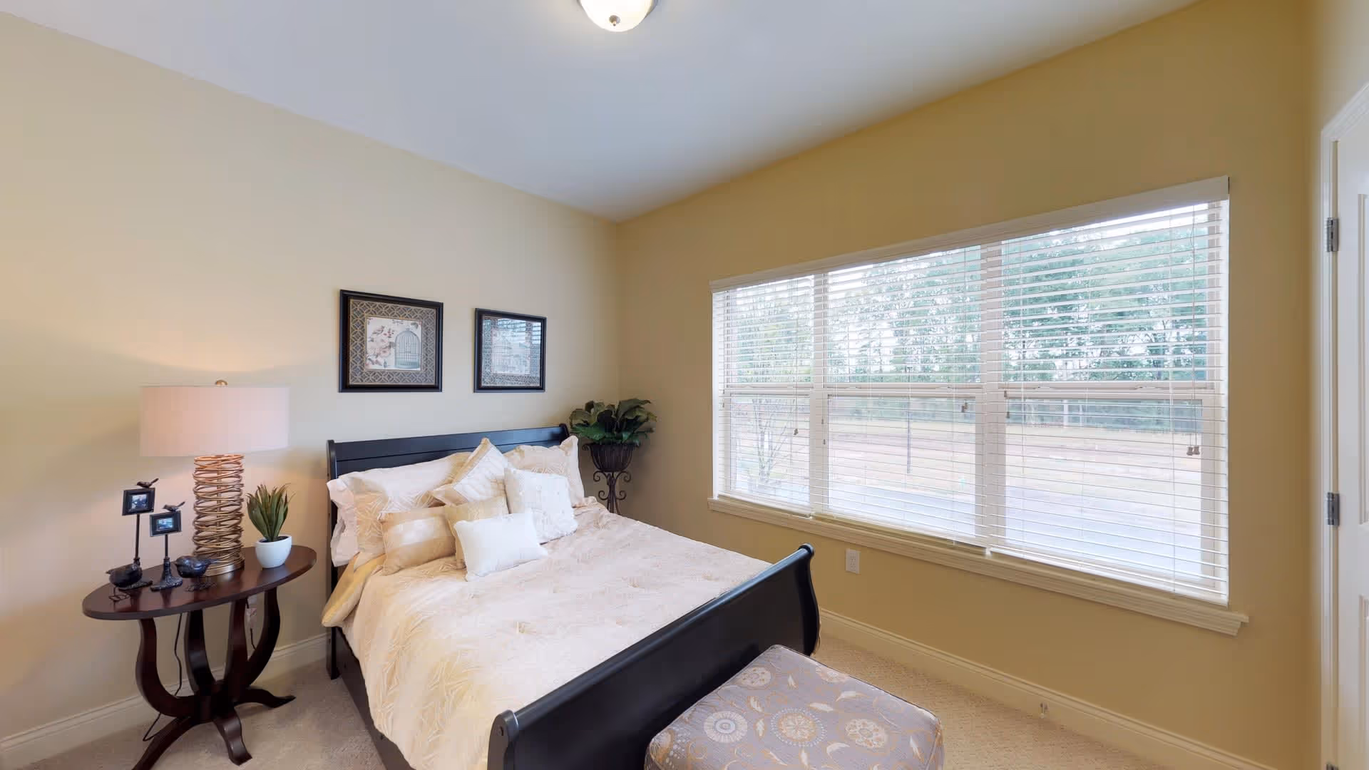 A cozy bedroom with a large window covered by white blinds letting in natural light. The room features a dark wooden bed frame with cream-colored bedding and multiple pillows. Next to the bed is a round wooden side table with a lamp, small plant, and decorative items. Two framed pictures hang on the wall above the bed, and a potted plant is placed in the corner near the window.