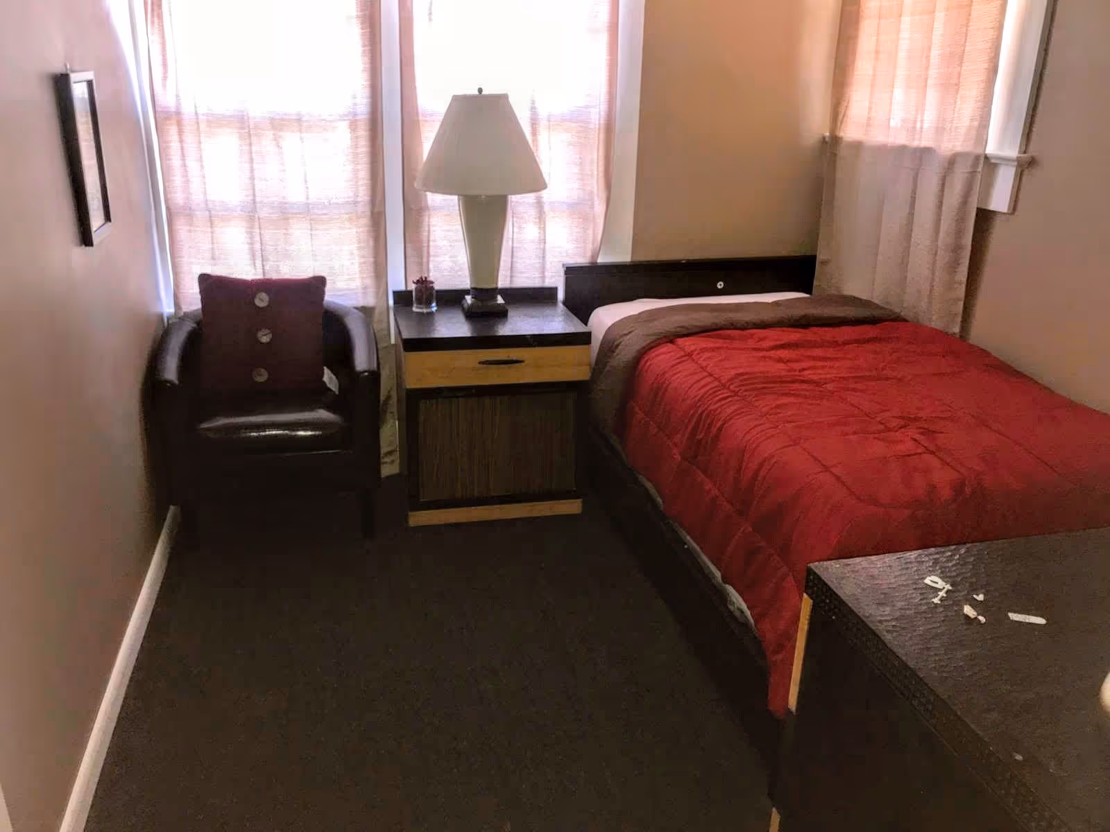 A small bedroom with a single bed covered with a red comforter, a nightstand with a lamp and a small plant, a dark brown armchair with a red cushion, and windows with light-colored curtains.