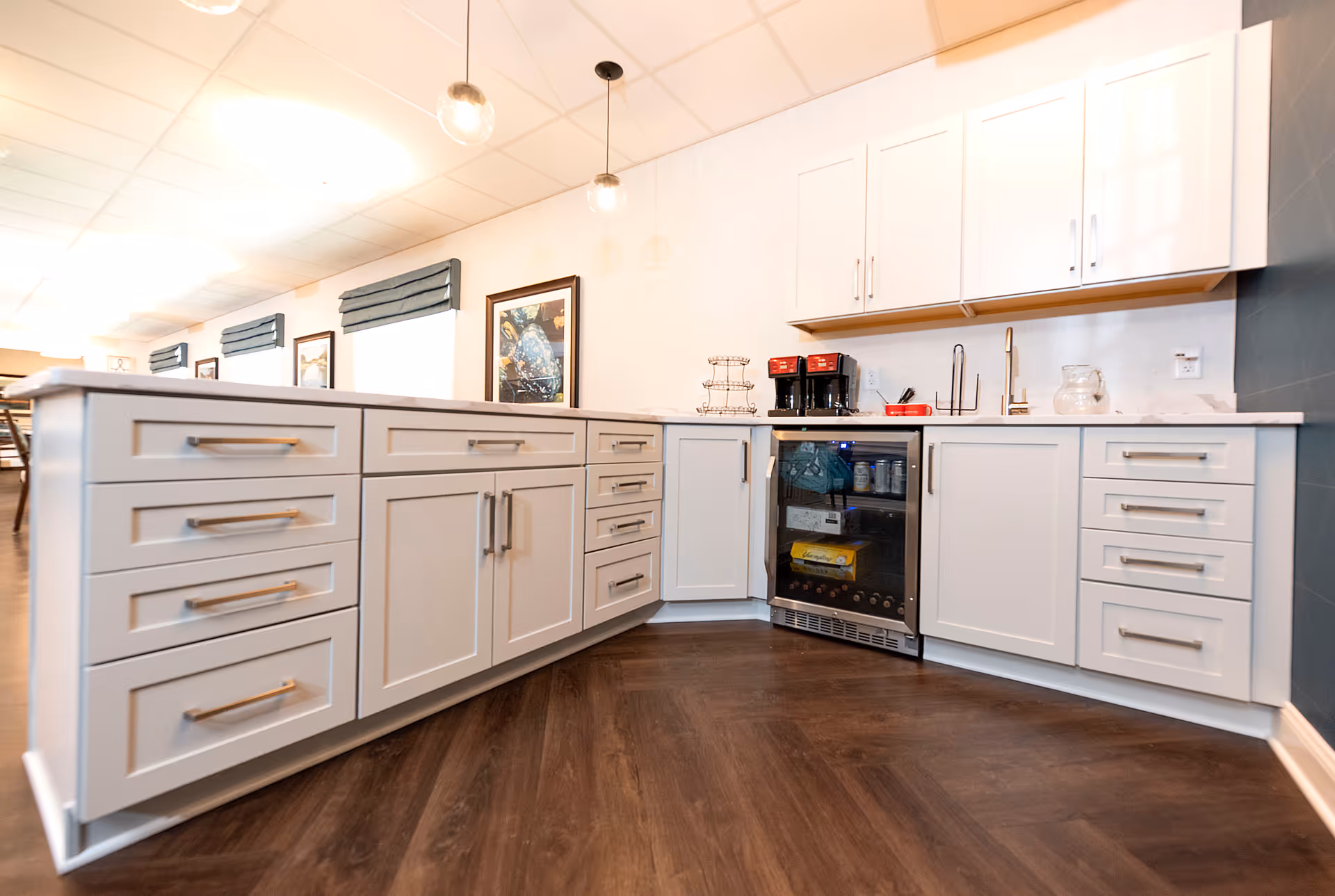 Modern kitchen area with white cabinets and drawers, a small built-in refrigerator with glass door, two coffee makers on the countertop, a sink, and pendant lights hanging from the ceiling. The floor is dark wood, and there are framed pictures and windows with dark blinds in the background.