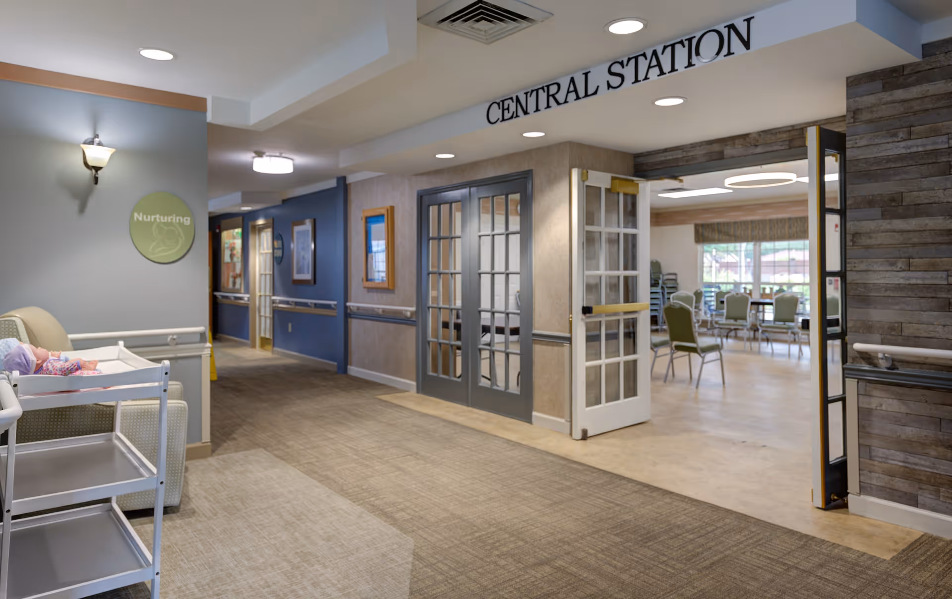 Interior hallway of Arden Courts A ProMedica Memory Care Community in Richardson, showing a carpeted corridor with handrails, a sign labeled 'Nurturing' on the wall, and an open double door leading to a room with chairs and tables. Above the door is a sign that reads 'CENTRAL STATION'.