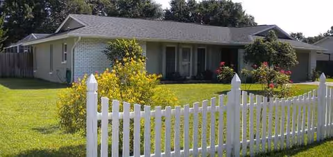 Exterior view of a single-story assisted living facility with a white picket fence and well-kept lawn.