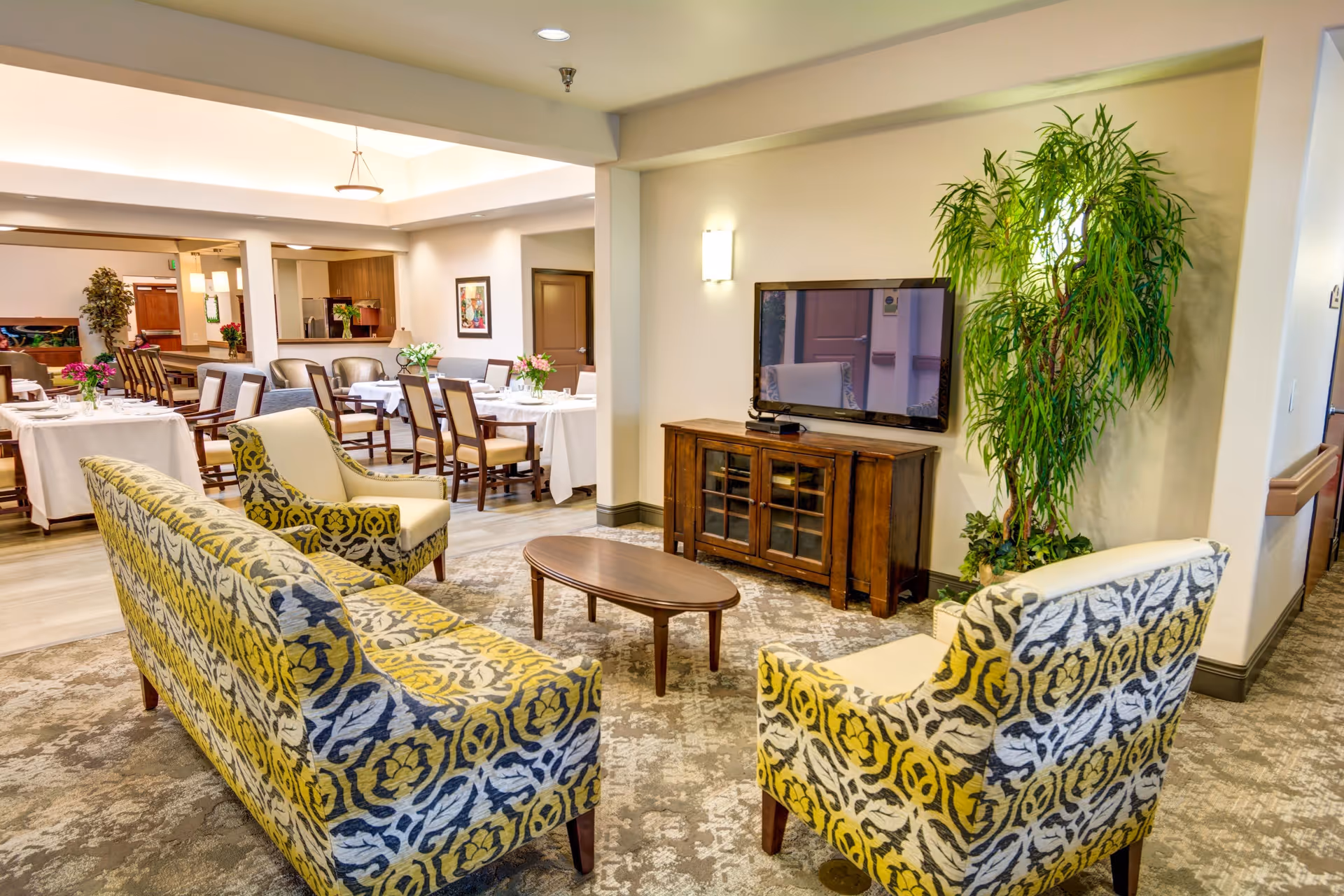 A cozy living area in a senior living facility with patterned yellow and gray upholstered chairs and sofa arranged around a wooden coffee table. A flat-screen TV sits on a wooden cabinet against the wall, and a tall green plant is placed beside it. In the background, there is a dining area with tables covered in white tablecloths and chairs neatly arranged, along with some floral centerpieces.