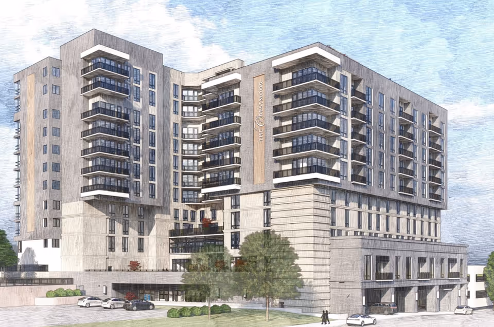 Exterior view of a modern multi-story senior living facility named The Crestmoor, featuring multiple balconies, large windows, and a landscaped area with trees and parked cars in front.