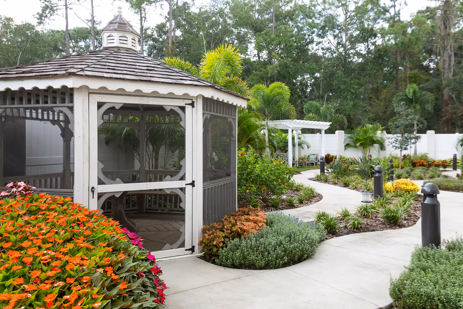A peaceful outdoor garden area featuring a white screened gazebo surrounded by colorful flowers and lush greenery. A curved concrete pathway winds through the garden, leading to a white pergola with seating. Tall trees and a white fence enclose the space.
