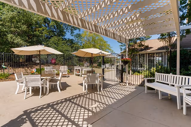 Outdoor patio area with white tables and chairs under large beige umbrellas, a white pergola casting patterned shadows on the concrete floor, surrounded by greenery and trees, with a building visible in the background.