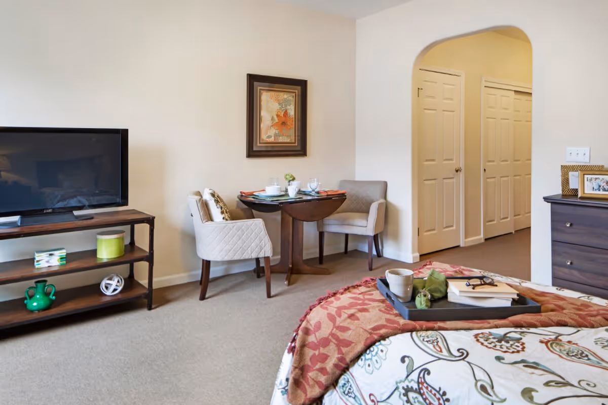 Well-lit bedroom with a bed in the foreground, a TV on a stand, a small round table with two chairs, and a dresser near an arched doorway and closet doors.
