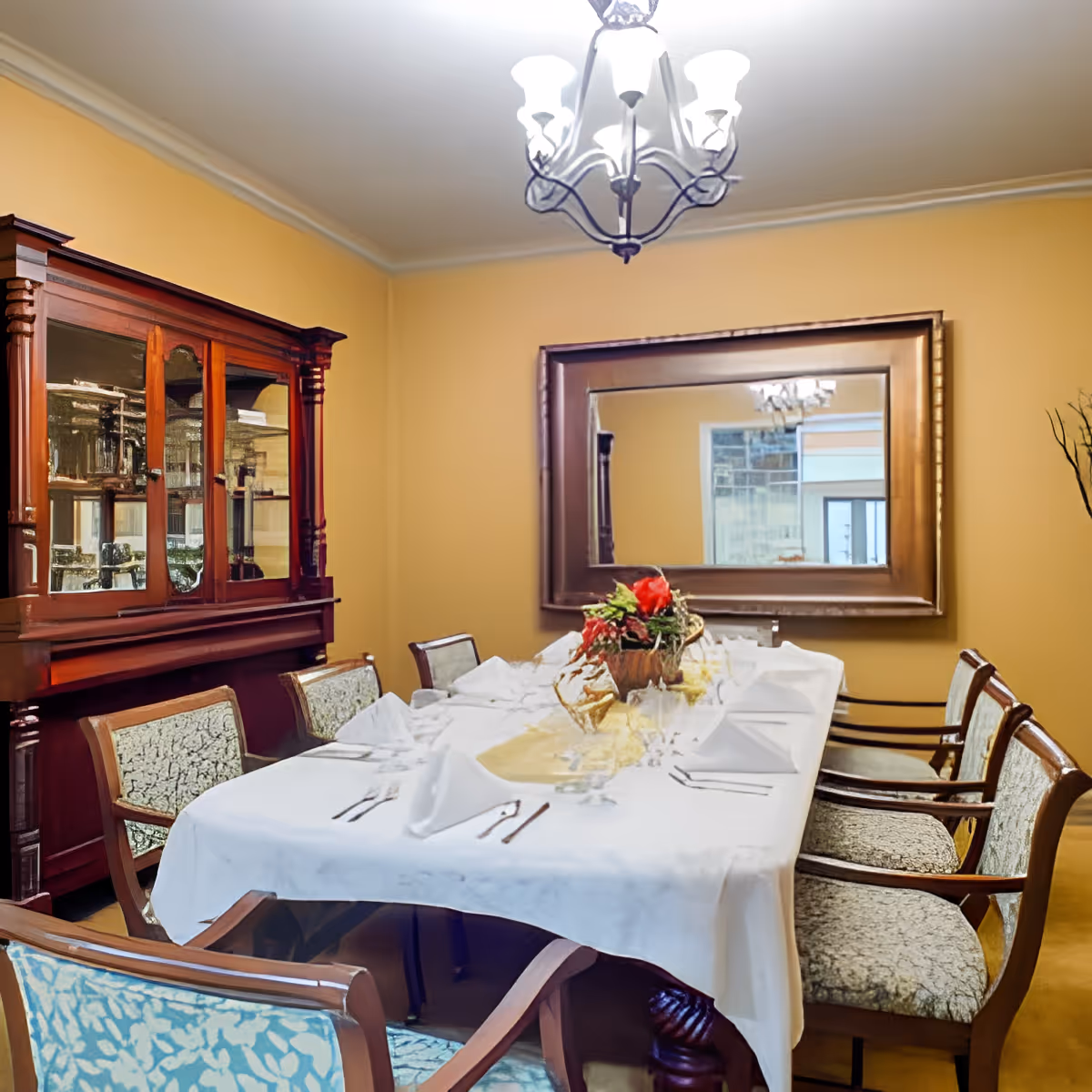 A formal dining room with a long table covered with a white tablecloth, set with folded napkins, glasses, and silverware. There is a floral centerpiece on the table. The room has upholstered wooden chairs around the table, a large wooden china cabinet with glass doors on the left, a large rectangular mirror on the wall, and a chandelier hanging from the ceiling.