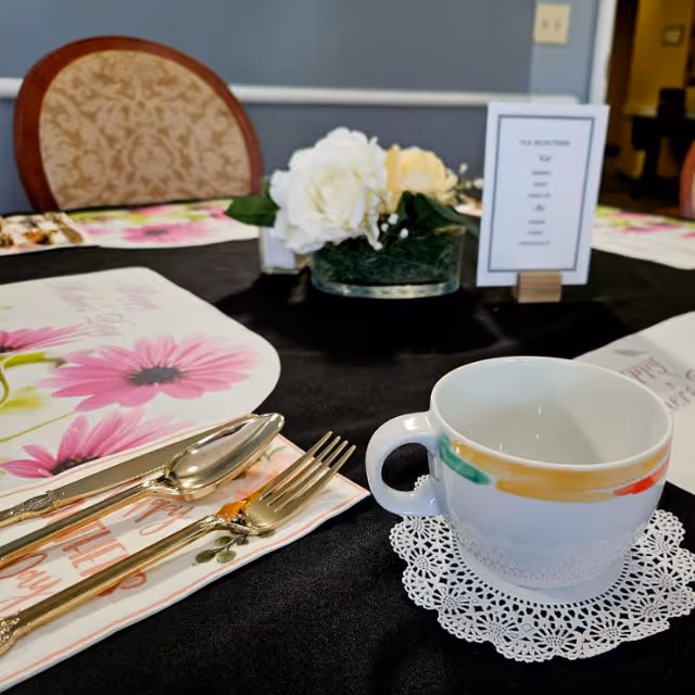 A dining table set with a teacup on a lace doily, gold flatware, floral placemats and a white flower centerpiece.