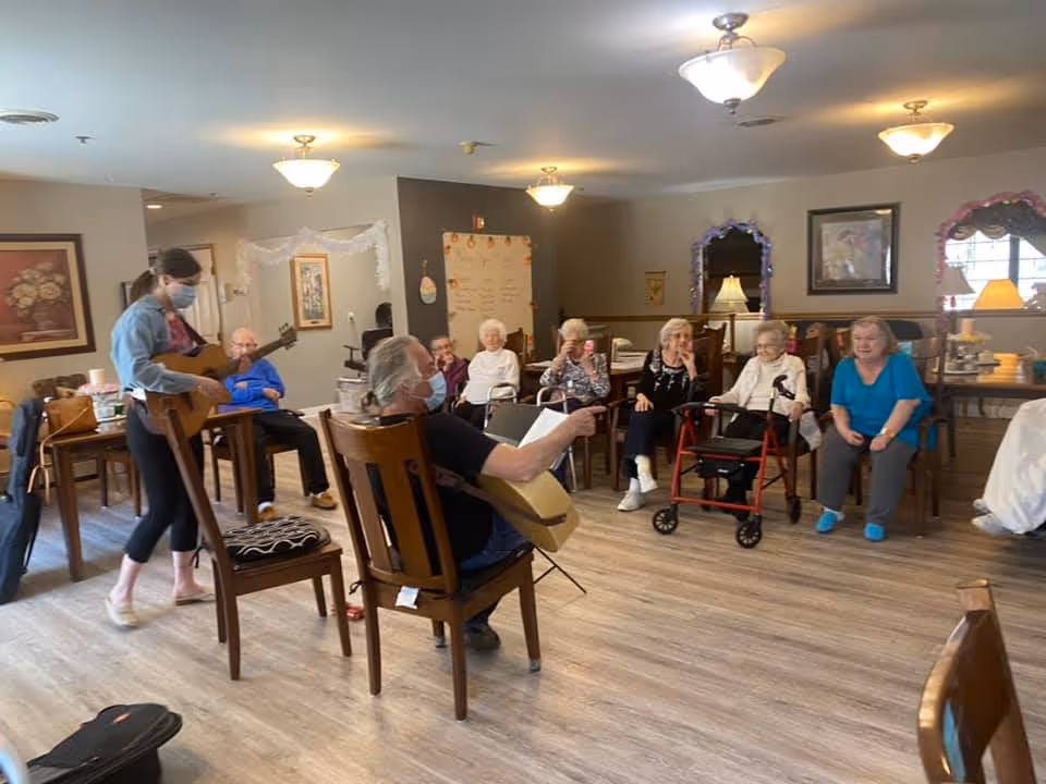 A group of elderly people seated in a semi-circle in a common room, watching two individuals playing guitars. The room has wooden flooring, several chairs, framed pictures on the walls, and ceiling lights. One guitar player is standing and wearing a face mask, while the other is seated and also wearing a mask.