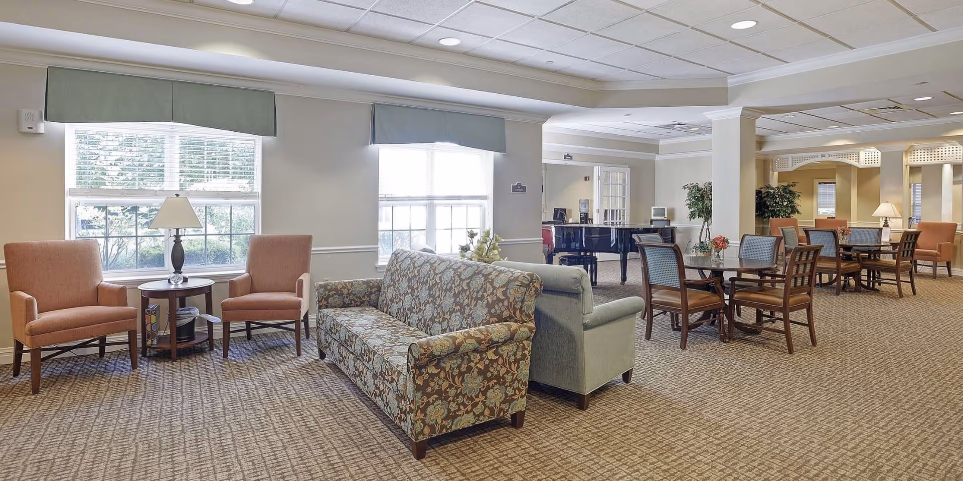 Bright communal living area with sofas, armchairs, dining tables, and a piano near large windows.