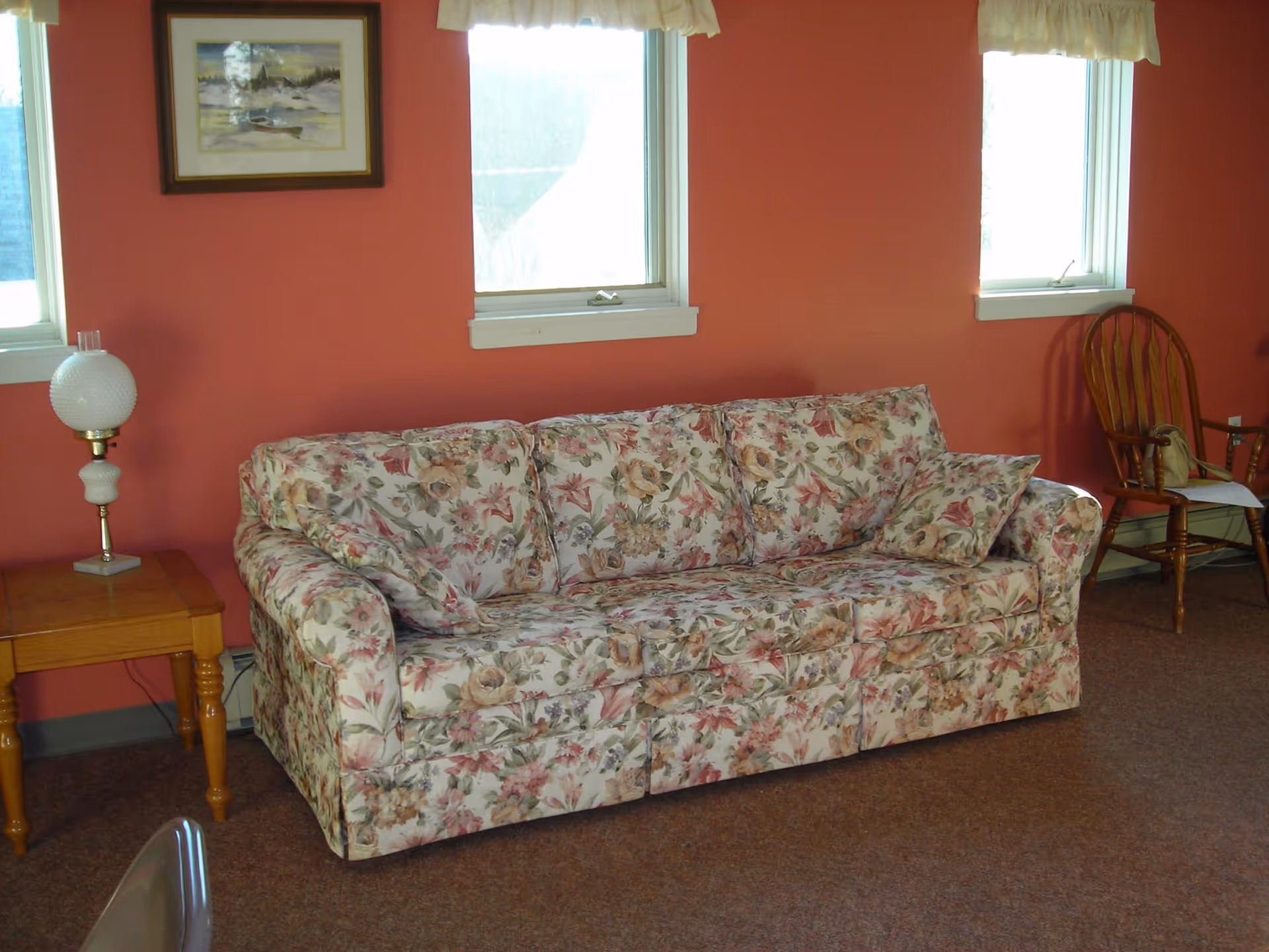 A floral patterned sofa with matching cushions is placed against a red wall with three windows above it. To the left of the sofa is a wooden side table with a vintage white lamp. To the right is a wooden rocking chair with a bag and some papers on it. A framed painting hangs on the wall above the sofa.