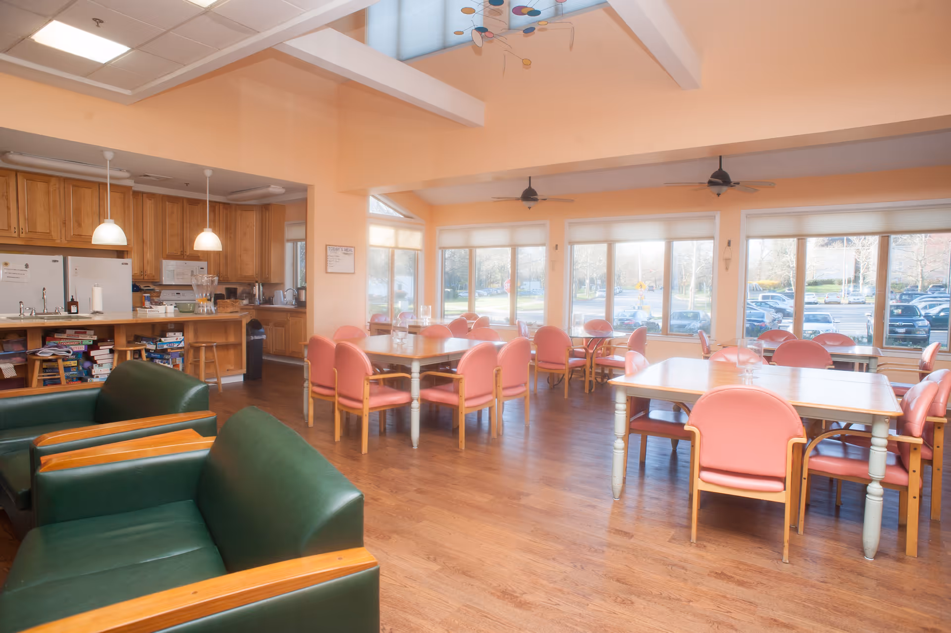 A bright and spacious dining area with several tables surrounded by pink cushioned chairs. The room has large windows letting in natural light and offering a view of a parking lot outside. There are green cushioned armchairs in the foreground and a kitchen area with wooden cabinets and white appliances in the background. The ceiling is vaulted with exposed beams and ceiling fans.